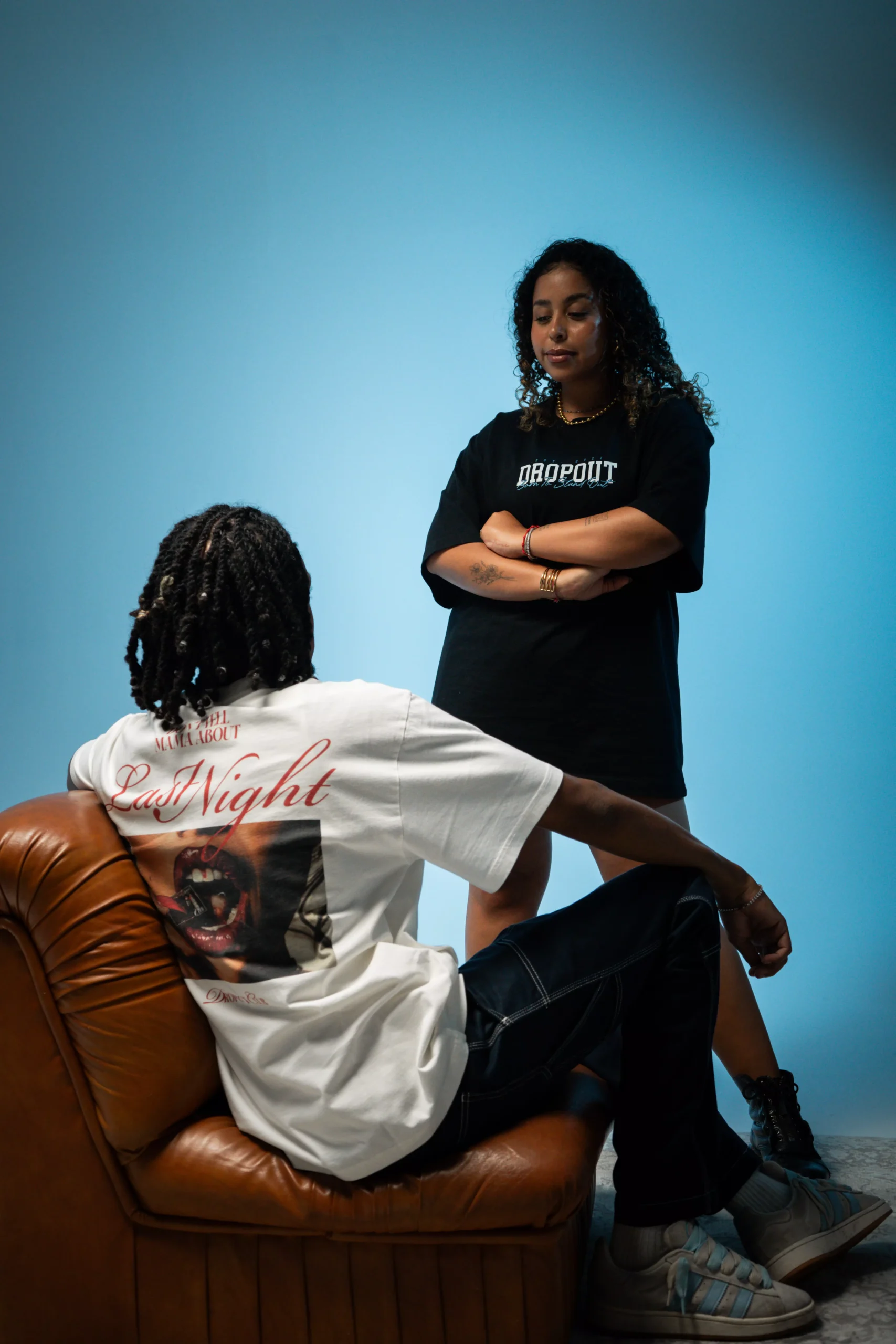 Flacko sits wearing the Hangover Oversized Black T-shirt while looking at Júlia who is wearing the Teddy Bear Streetwear T-shirt from the Dropout brand.