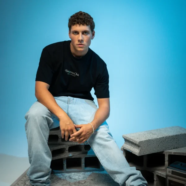 Gustavo wearing the Tasty Poison Black T-shirt by Dropout while sitting on bricks with one leg stretched out