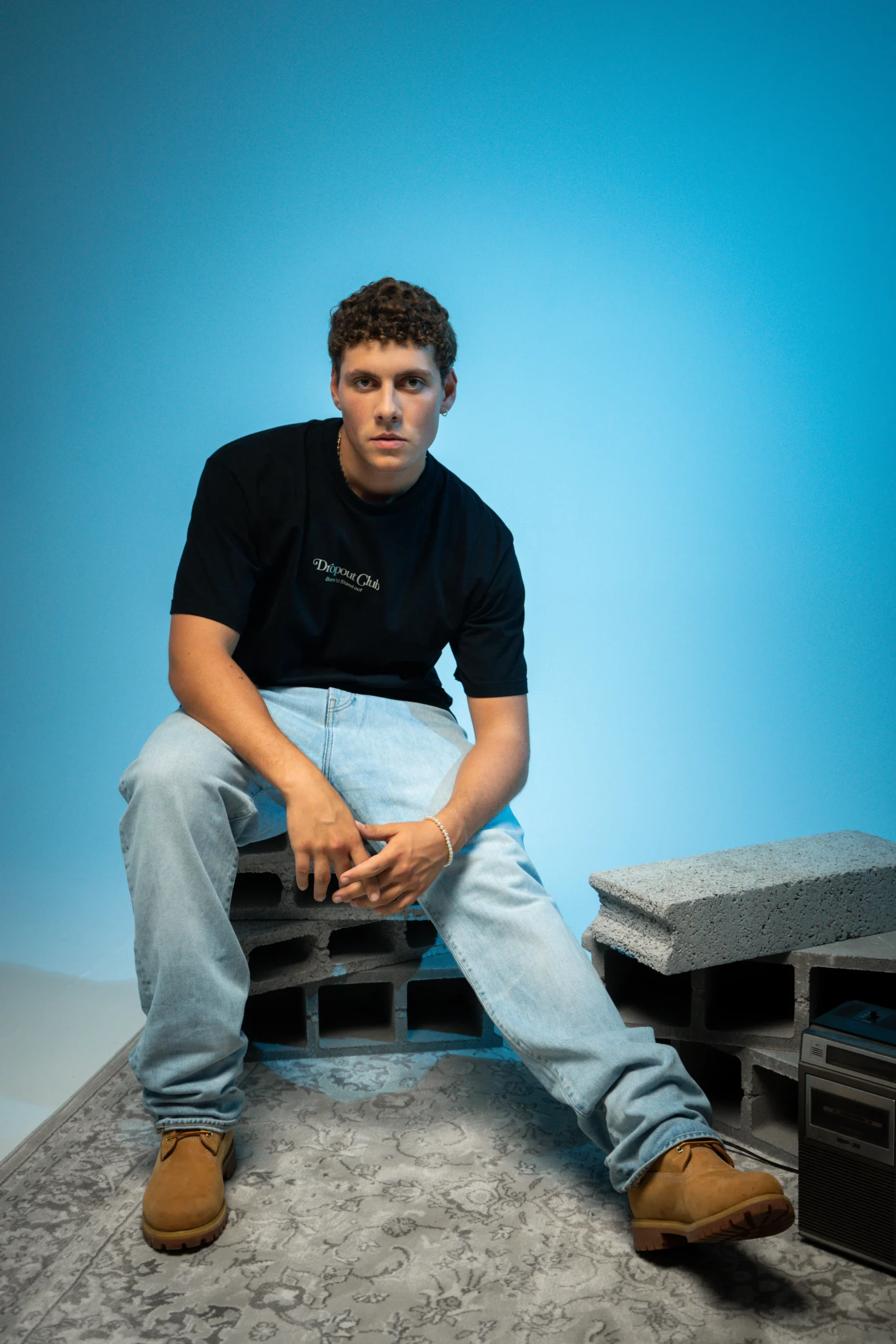 Gustavo wearing the Tasty Poison Black T-shirt by Dropout while sitting on bricks with one leg stretched out